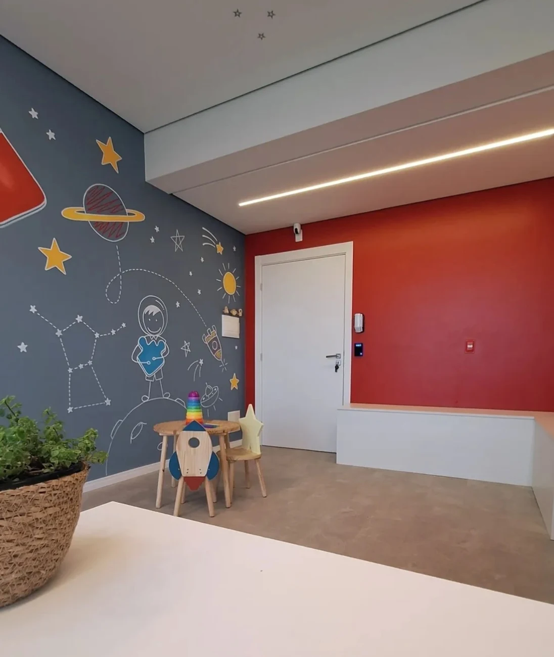 Foto real do consultório com mesa infantil, cadeiras de foguete e estrela e mural de planetas.